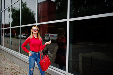 Portrait of a fabulous young woman in red blouse and jeans posing with her handbag and sunglasses outside the shopping mall on glass background.