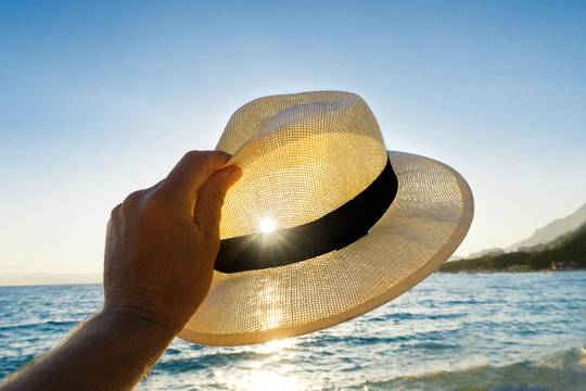 Hand Holding A Straw Hat Against The Sun And Sea.