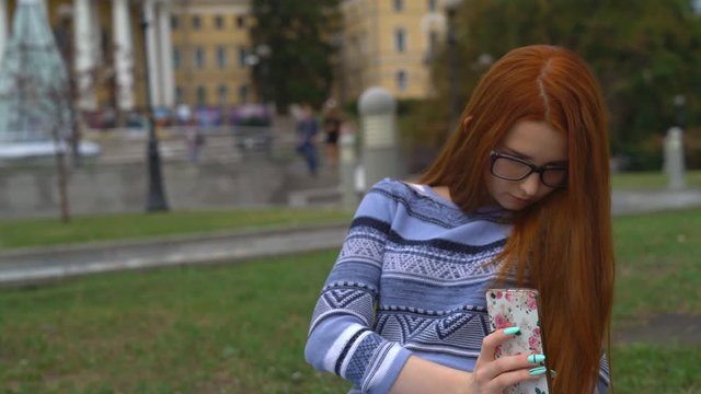 A young teenage girl with red hair and blue eyes is sitting on the parapet and taking pictures on her mobile phone.