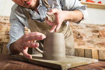 pottery, stoneware, ceramics art concept - young male stands at a workshop behind the table. workplace of craftsman with an unfinished product of clay cup and connected handle, front view