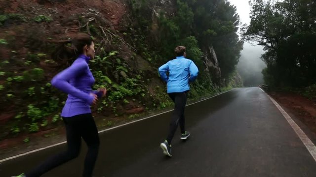 Fitness Sport Couple Running. Tracking shot of multiethnic couple Jogging on wet country road. Determined man and woman are jogging by mountains. They are representing their healthy lifestyle.