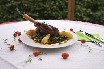 Slowly roasted lamb knuckle with thyme, pepper and garlic served with green spinach and boiled new potatoes as a side dish. Meal is on the white rustic plate on the table in garden restaurant.