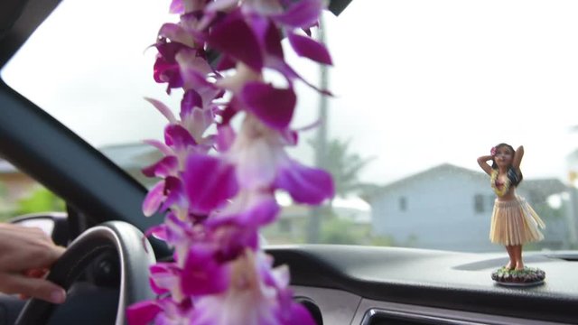 Closeup of hula doll on dashboard with orchid garland hanging in foreground. Cropped image of hand is driving car. Figurine is dancing in vehicle.