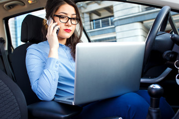 Beautiful young business woman sitting in the car with laptop and talking on phone.