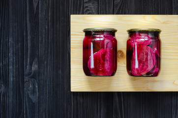 Canned homemade beetroot in two clear glass jars with wooden cutting board on a black wooden background with copy space for your text message or promotional content.