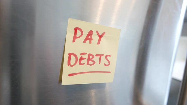 Putting a PAY DEBTS sticky note reminder on a fridge. Closeup on the hand and paper.
