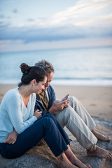 Fototapeta na wymiar A couple sitting at the beach, looking at photos on a phone