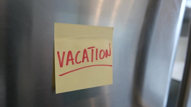 Putting a Vacation sticky note reminder on a fridge. Closeup on the hand and paper.