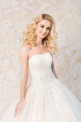 Fototapeta na wymiar fashionable white gown, beautiful blonde model, bride hairstyle and makeup concept - young smiling girl in wedding festive dress, standing indoors on light background, romantic slender woman posing