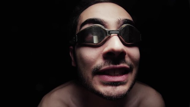A weird looking man close up wearing googles and making signs with his hands. Pure black background.