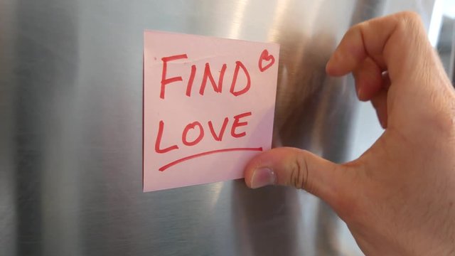 Putting a Find Love sticky note reminder on a fridge. Closeup on the hand and paper.