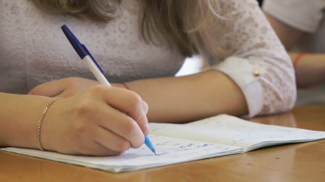 Student dressed in a white shirt sits at a school desk writing text in exercise book using ballpoint pen. Close-up