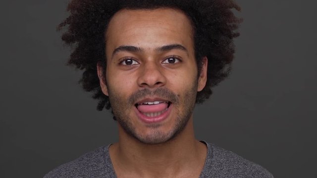 Dark skinned young man with afro haircut having fun showing his white teeth ang tongue over charcoal background closeup. Concept of emotions