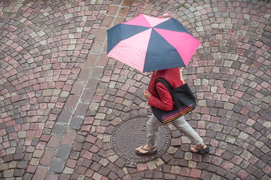 femme avec parapluie traversant une place pavés sous la pluie