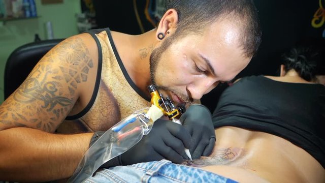 Portrait of man tattoo master with dreadlocks showing process of creation tattoo on female body under the lamp light. Slow motion. Professional artist working in salon, close up view.