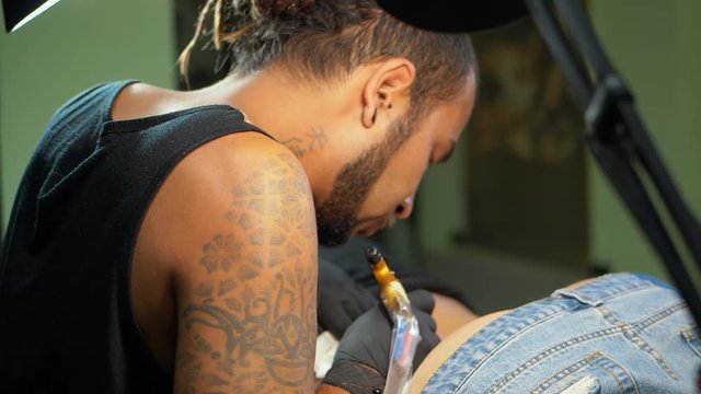 Tattooist does tattoo a client with machine close up. Process getting tattoo in a parlor. Body tattooing process. Man with dreadlocks working on female body part under lamp. Slow motion.