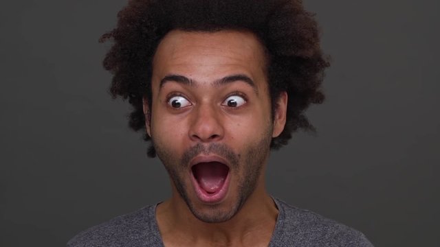 Dark skinned handsome man joyfully surprised with open mouth, isolated on charcoal background closeup. Concept of emotions