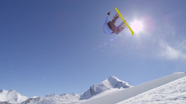 SLOW MOTION: Young pro snowboarder riding the half pipe in big mountain snow park, jumping out of the halfpipe wall and over the sun, performing tricks and rotations with grabs in sunny winter 