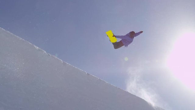 SLOW MOTION: Young pro snowboarder riding the half pipe in big mountain snow park, jumping out of the halfpipe wall and over the sun, performing tricks and rotations with grabs in sunny winter 