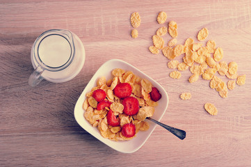 cornflakes with strawberries and a jug of milk on wooden background