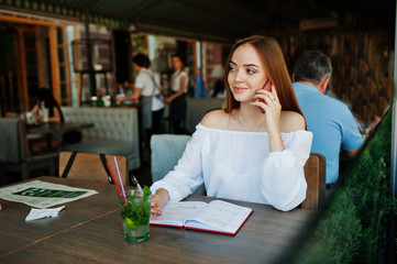 Portrait of a gorgeous young businesswoman talking on the phone and writing something down in her red notebook while sitting in a cafe.