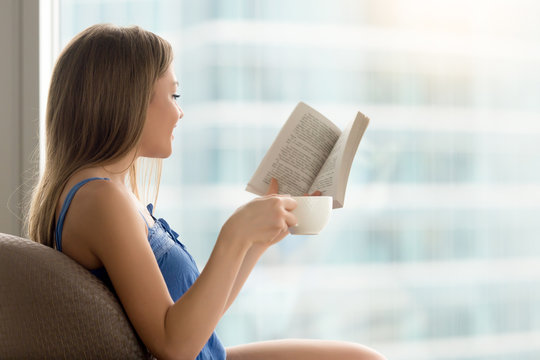 Young casual woman sitting in chair, reading interesting paper book, and drinking coffee. Lady resting at comfort home with favorite book in hand, enjoying good literature, relaxing in cafe on weekend