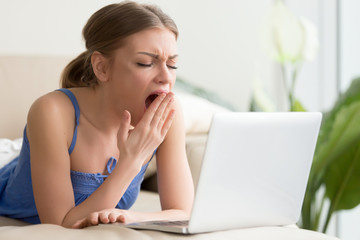 Bored young woman lying on sofa in front of laptop and yawning. Attractive lady boring because of...