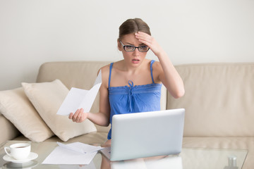 Worried young woman in eyeglasses sitting on sofa with paper letter in hand and looking with confusion on laptop screen. Female taxpayer with overdue tax returns shocked because of financial penalties