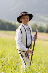 Child boy in traditional goral dress, hat and axe stands on meadow in Polish mountains