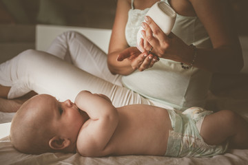 Mother with her baby at home. Mother uses a body cream, and putting of a baby.