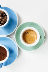 Three cups of coffee: ground coffee (powder), coffee beans and freshly made espresso in colorful ceramic cups (mint green and light blue) on the white background