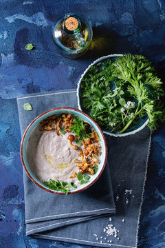 Mushroom cream soup in ceramic bowl served with forest mushrooms, greens, fried onion, salt, bottle of olive oil on blue textile napkin over dark blue texture concrete background. Top view
