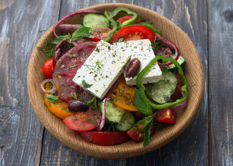 Homemade Greek salad from tomatoes, cucumbers, sweet peppers, cheese, olives and greens in a wooden bowl on a wooden table. Healthy delicious food