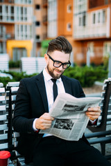Handsome bearded businessman in classic suit is drinking coffee and reading a newspaper while resting on the bench in the city