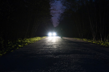 Male silhouette at the edge of a dark mountain road through the forest in the night. Man standing on the road against the car headlights. The car on the roadside. Mystery concept. Soft focus, filter.