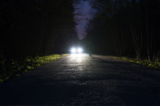 Male Silhouette At The Edge Of A Dark Mountain Road Through The Forest In The Night. Man Standing On The Road Against The Car Headlights. The Car On The Roadside. Mystery Concept. Soft Focus, Filter.