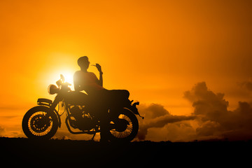 Silhouette of biker man sitting smokes with his motorbike beside the natural lake and beautiful, enjoying freedom and active lifestyle, having fun on a bikers tour.sunset background and sky.