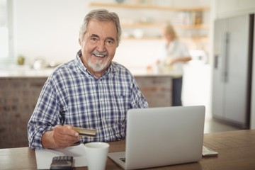 Smiling senior man paying bills online on laptop in kitchen