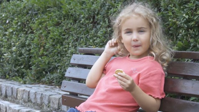 A little girl is sitting on a bench and eating biscuit. Сhild has a snack in the park outdoors.