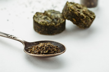 Fototapeta na wymiar Heap of dry green tea in a metal spoon and compressed tea in the background