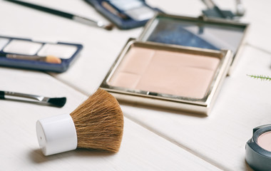 Cosmetic brush with open powder box among other cosmetics on a white wooden table