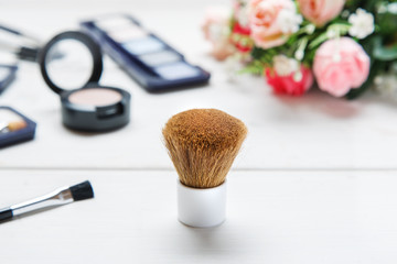 Obraz na płótnie Canvas Big cosmetic brush among makeup items on a white wooden table with roses bouquet on the background, closeup shot, selective focus