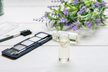 Cosmetic makeup items with open glass vial with liquid in it and lavender flower on a white wooden table