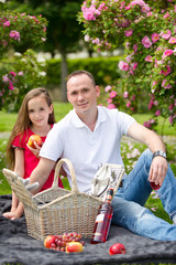 Beautiful young father sitting on a plaid in a green park with her small pretty daughter with a wicker basket for happy picnic
