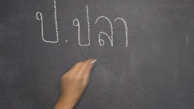 Woman's hand writing Thai letter "ก" with white chalk on blackboard