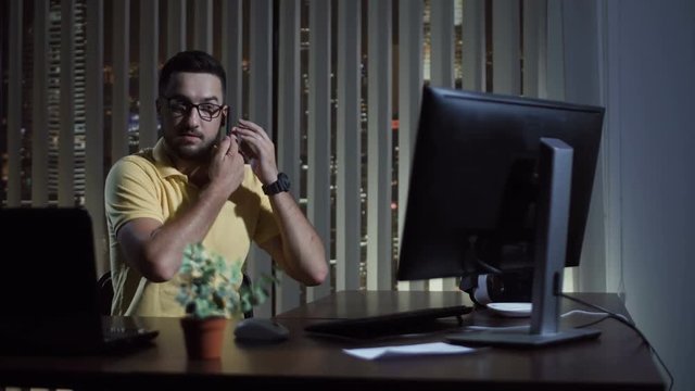 Man in casual wear working in office at night sitting at laptop and communicating with phone.