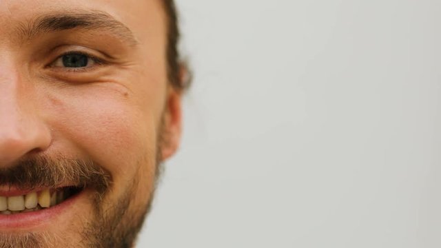 Portrait of half face of attractive man with beard smiling on camera on the white wall background. Cropped view