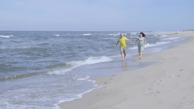 Happy lovely couple running along the seacoast barefoot and enjoys herself. Beach with white sand. Outgoing coastal line. Slow Motion.