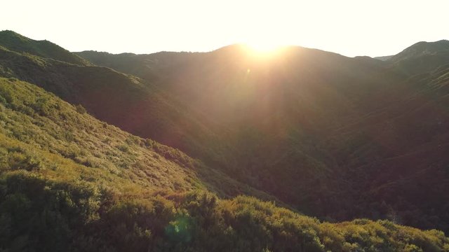 Beautiful aerial sunset shot flying forwards over forested hills. Flies over a ridge revealing valley below. Lush green trees and dazzling sunset. Lens flare.