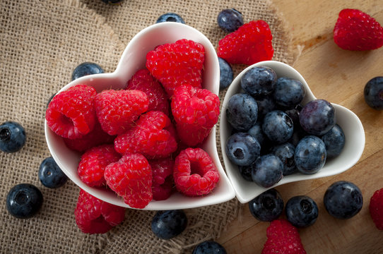 Heart healthy foods and good eating habits concept with two heart shaped white bowls of blueberries and raspberries with copy space. The blueberry bowl is smaller than the raspberry one